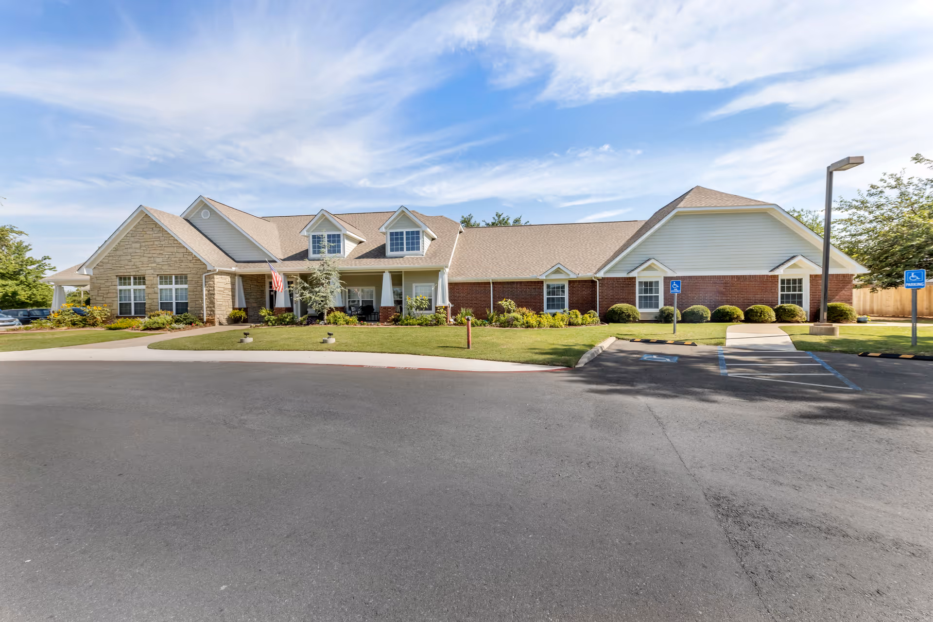Single-story brick and siding senior living building with a front porch, American flag, parking spaces, and landscaped lawn under a blue sky.