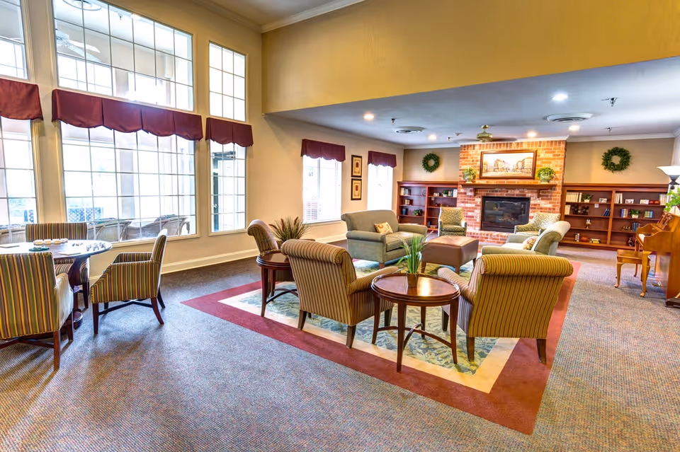 A cozy living room area with multiple upholstered armchairs and sofas arranged around a central ottoman and a fireplace with a brick surround. Large windows with red valances allow natural light to fill the room. There are bookshelves on either side of the fireplace, decorative wreaths on the walls, and a piano in the corner.