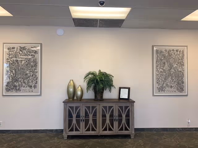 A beige wall with two framed black and white botanical prints hanging on either side of a wooden cabinet. On top of the cabinet are two gold vases, a green potted plant, and a small framed sign. The floor is carpeted with a patterned design, and the ceiling has fluorescent lights.