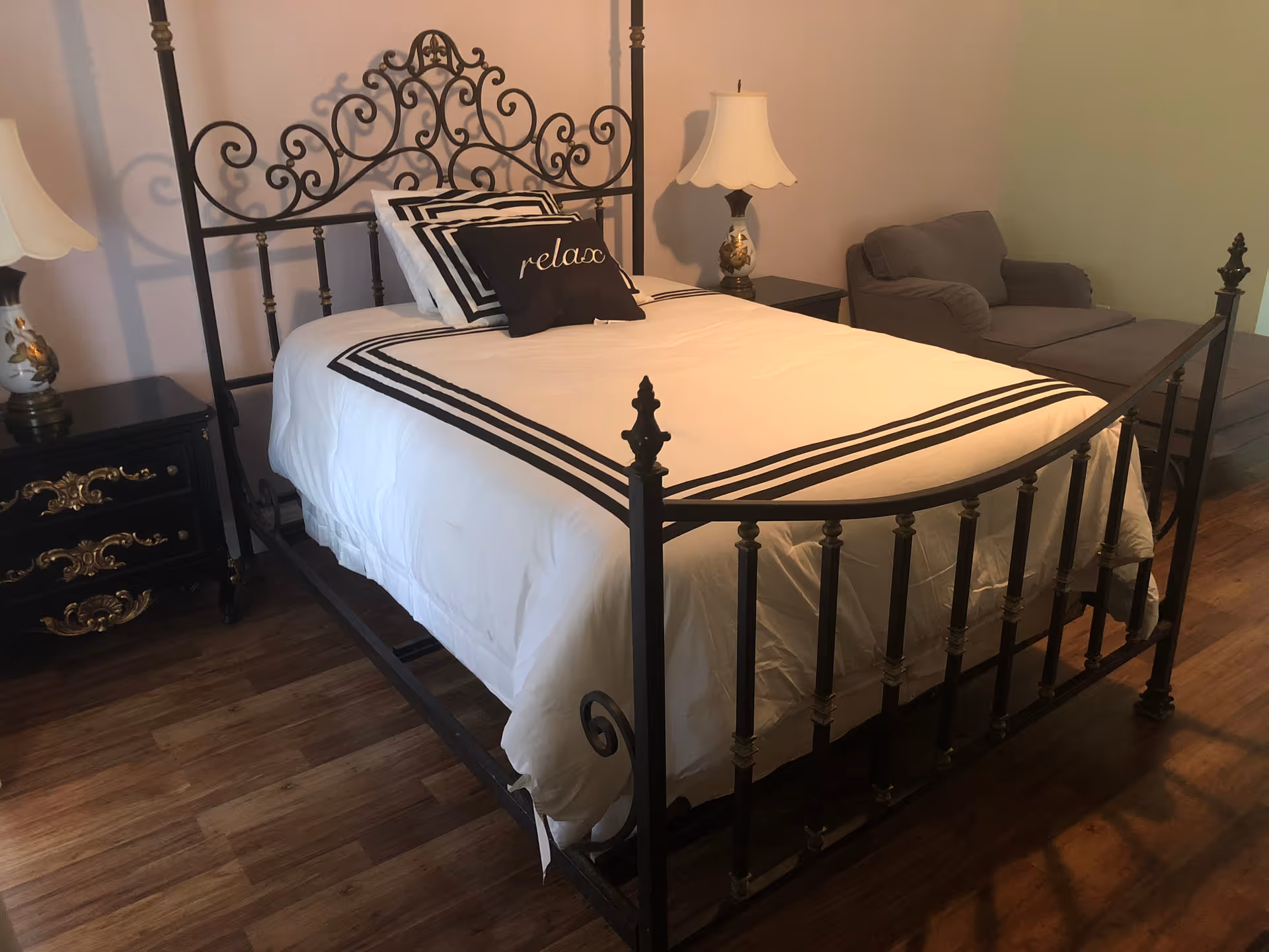 A bedroom with a decorative black metal bed frame, white bedding with black stripes, and a black pillow with the word 'relax' on it. There are two bedside tables with matching lamps, and a gray armchair is positioned in the corner of the room. The floor is wooden.