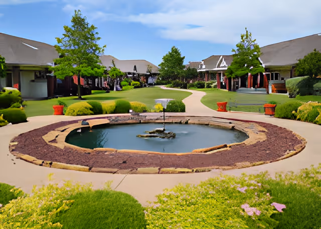 A landscaped outdoor courtyard with a circular water fountain in the center, surrounded by a paved walkway. The courtyard is bordered by green bushes, trees, and buildings with sloped roofs under a partly cloudy sky.