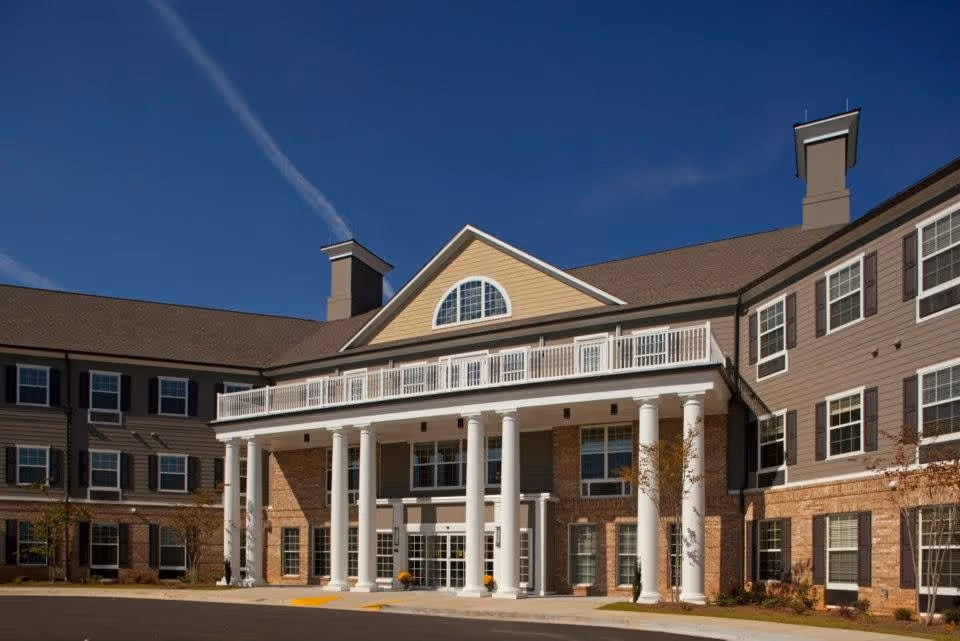 Exterior view of a senior living facility building with a large covered entrance supported by white columns, multiple windows with shutters, and a clear blue sky in the background.