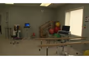 A physical therapy or rehabilitation room with parallel bars for walking exercises, exercise balls stacked in the corner, a computer on a rolling desk, and various medical equipment against the walls. The room has a window with a blind partially drawn and light-colored walls and flooring.