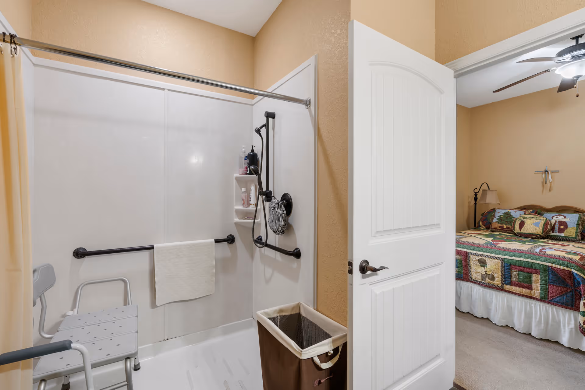 Accessible bathroom with a walk-in shower, grab bars, shower chair, and an open door leading to a bedroom.