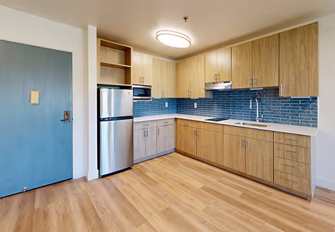 Modern kitchen with light wood cabinets, a stainless steel refrigerator, microwave, and range hood. The backsplash is made of blue rectangular tiles, and the floor is light wood. A blue door is visible on the left side of the image.