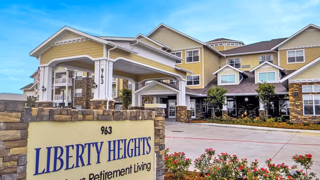 Exterior view of Liberty Heights Gracious Retirement Living facility showing a large multi-story building with beige and brown siding, stone accents, and a covered entrance. There is a sign in the foreground with the facility name and address number 963, surrounded by landscaping with flowers and shrubs under a blue sky.