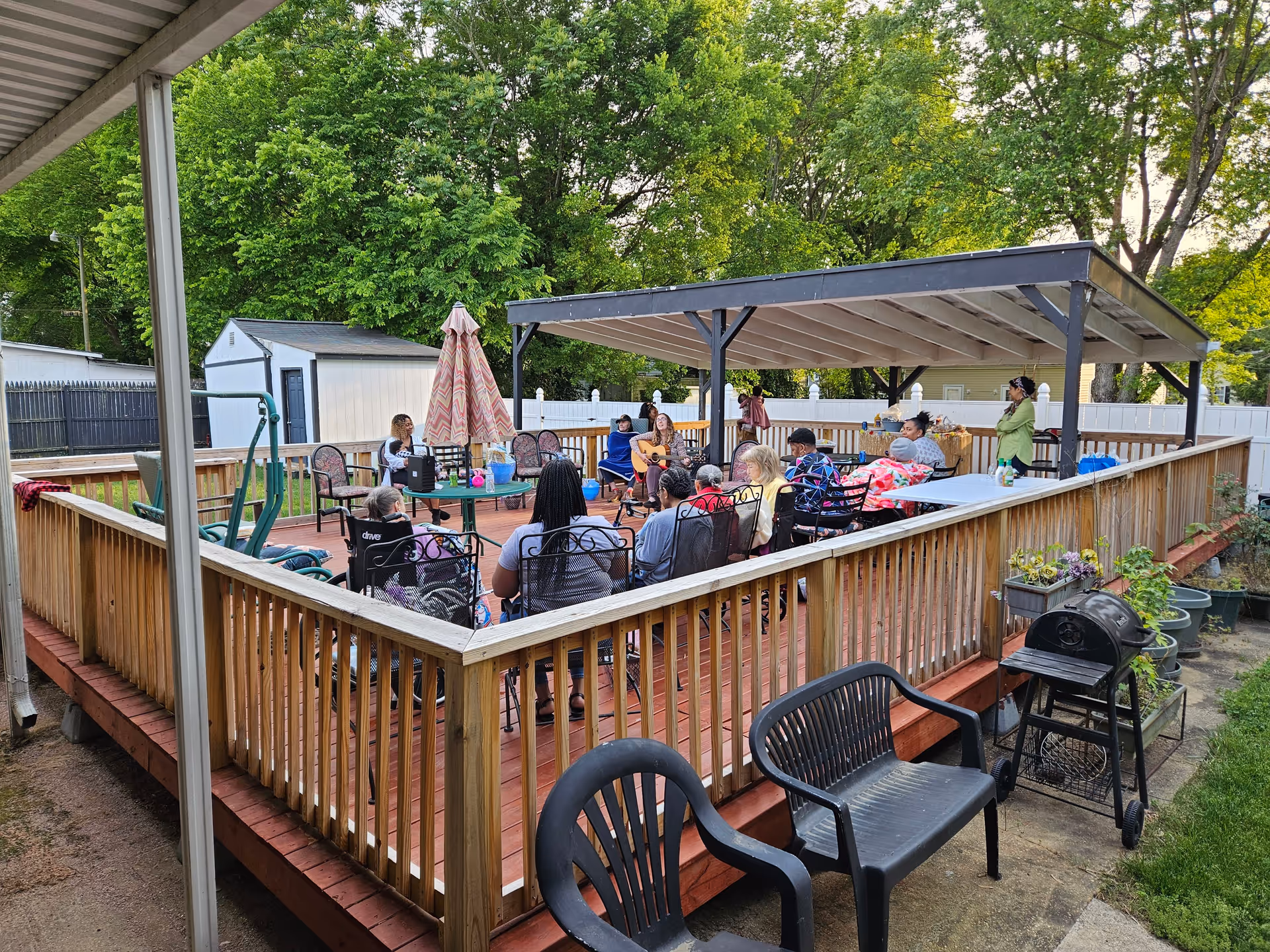 Outdoor wooden deck area with a group of people seated in chairs arranged in a circle under a covered pergola. There is a woman playing guitar and others listening. The deck is surrounded by a wooden railing and there are trees and greenery in the background. Plastic chairs and a small grill are visible outside the deck area.
