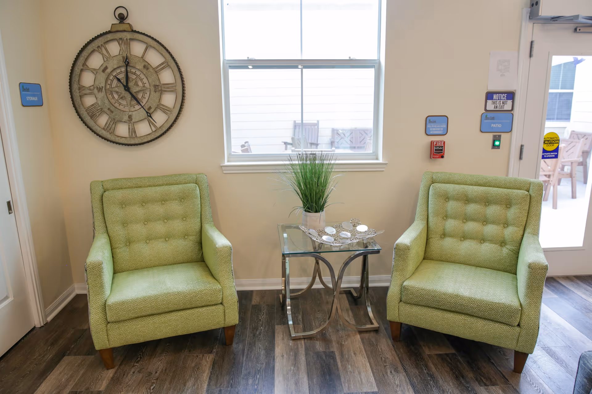 Two green upholstered armchairs flank a glass side table with a potted plant beneath a window and a decorative wall clock on the wall.