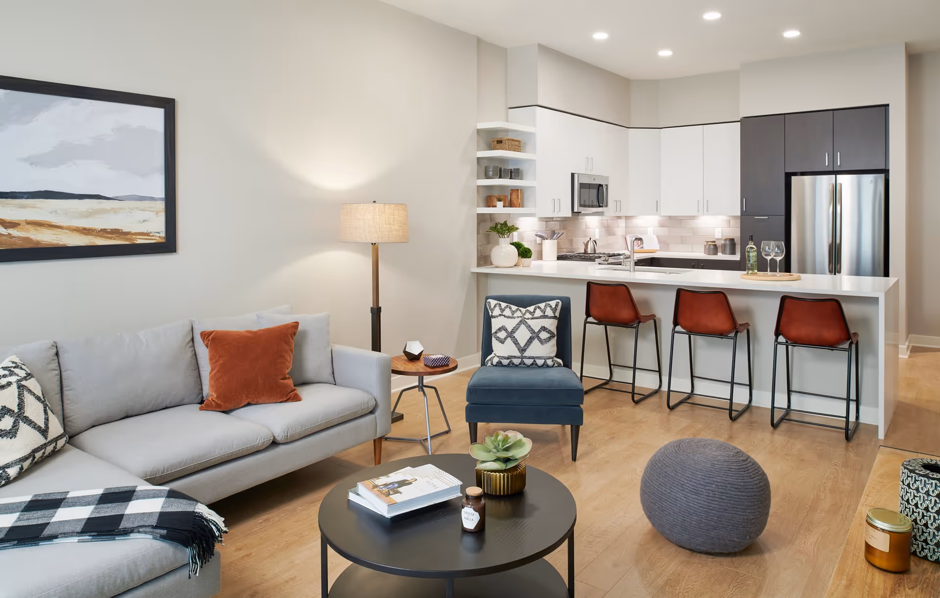 A modern open-concept living room and kitchen area featuring a light gray sectional sofa with patterned and rust-colored pillows, a round black coffee table with books and a small plant, a blue accent chair with a decorative pillow, a floor lamp, and a kitchen with white and dark cabinetry, stainless steel appliances, a white countertop island with three brown bar stools, and wooden flooring.
