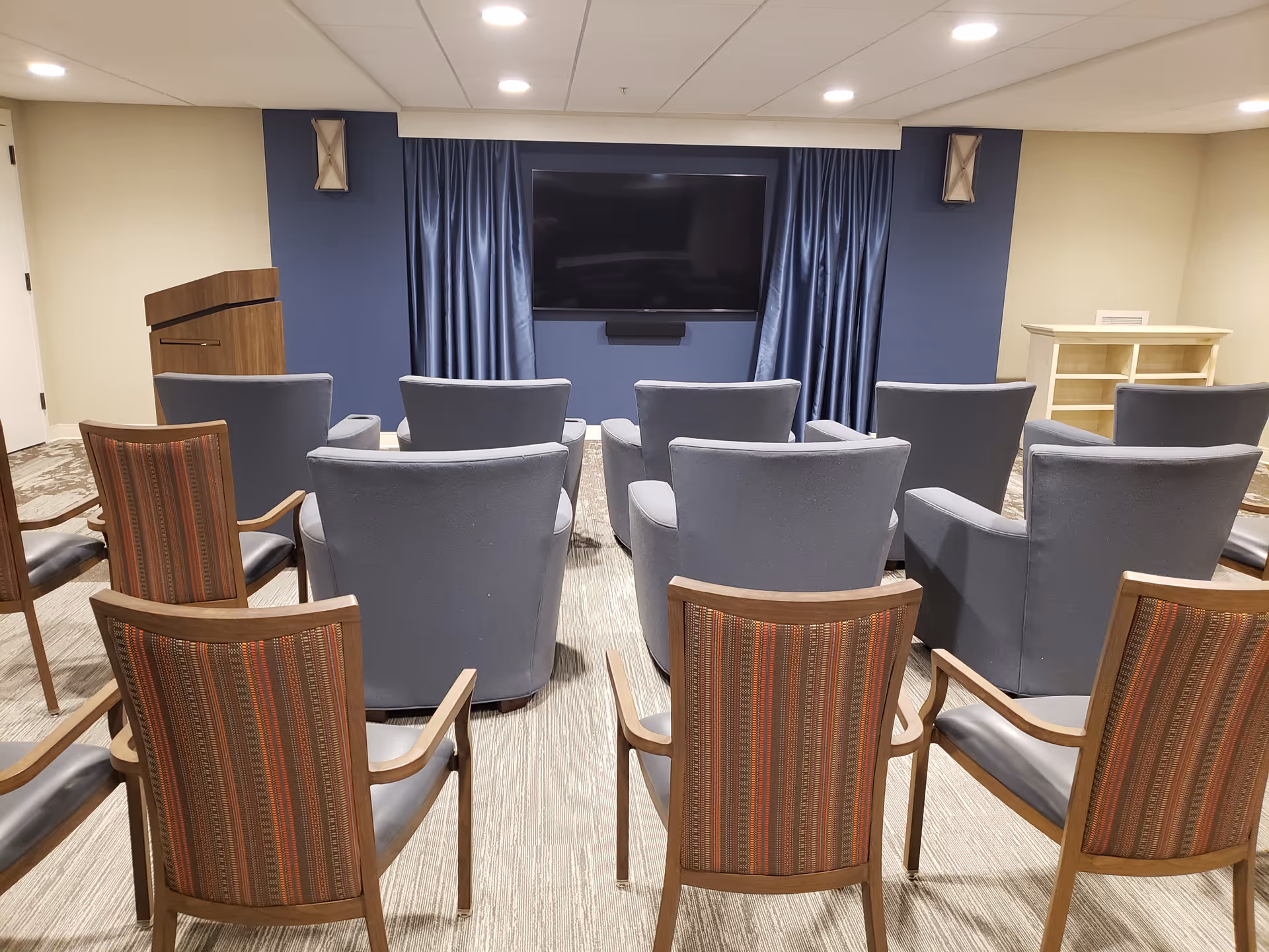 A small conference or media room with several rows of chairs facing a large flat-screen TV mounted on a blue accent wall with blue curtains on either side. The room has a podium on the left side and a small wooden shelving unit on the right. The floor is carpeted and the walls are painted beige.