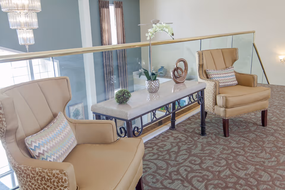 Two beige upholstered armchairs with patterned cushions are placed on a carpeted floor with a decorative design. Between the chairs is a rectangular table with a stone top and black metal legs, adorned with a small green plant, a white orchid in a silver pot, and a modern abstract sculpture. The area is next to a glass railing overlooking a lower level with large windows and curtains.