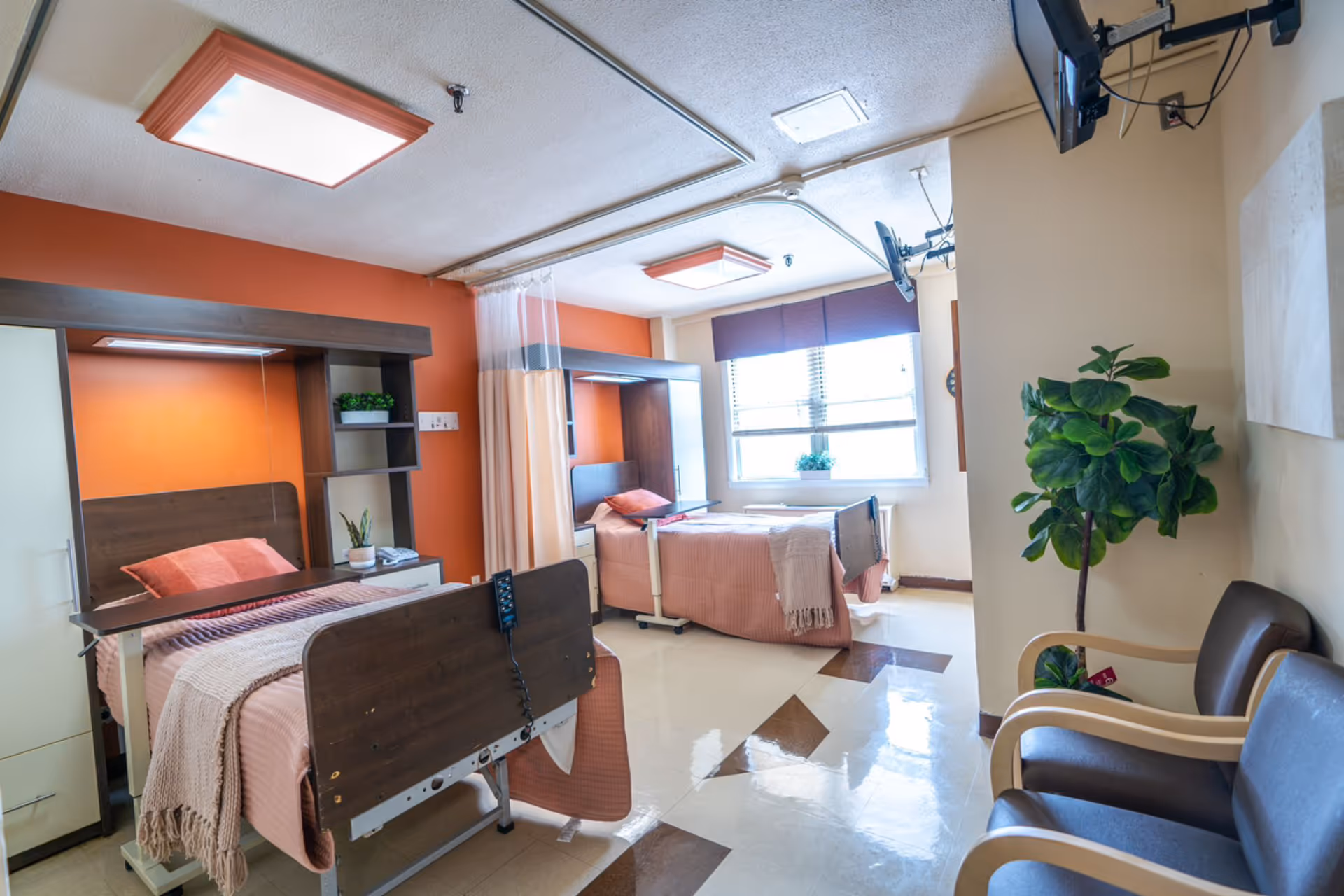 A bright nursing facility room with two hospital beds, each with a pink blanket and pillow. The beds are separated by a privacy curtain. The room has orange accent walls, a window with blinds, two wall-mounted televisions, a potted plant, and two chairs near the entrance.