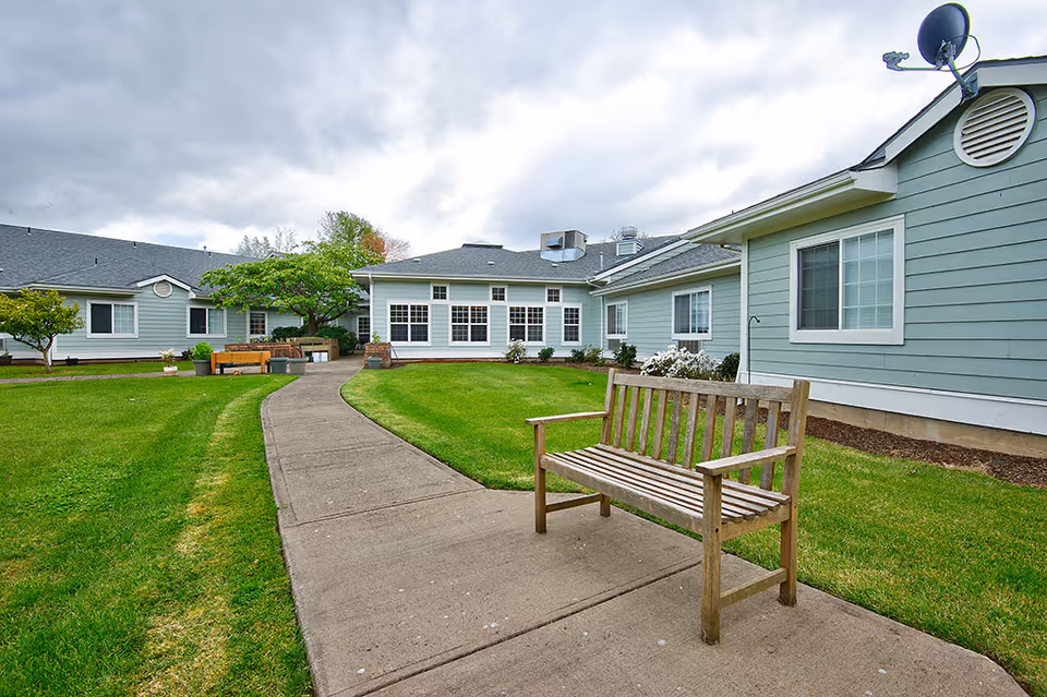 Outdoor view of Spring Valley Assisted Living facility showing a paved walkway through a green lawn with a wooden bench on the right side. The building has light blue siding with white trim and multiple windows. Trees and shrubs are visible around the building under a cloudy sky.