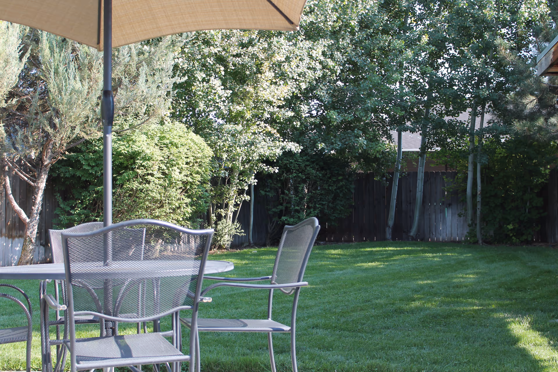 Outdoor patio area with metal mesh chairs and a table under a large beige umbrella, surrounded by green grass, trees, and bushes with a wooden fence in the background.