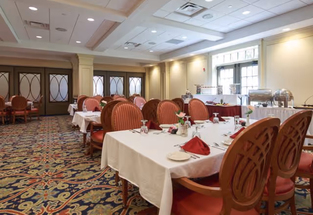 Well-appointed dining room with white-tablecloth tables, red upholstered chairs, patterned carpet, and a buffet setup along the far wall.