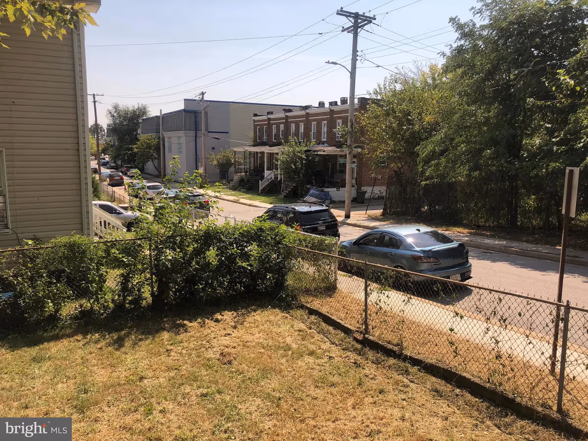 View of a residential street with parked cars, a chain-link fence, some bushes, and trees on a sunny day.