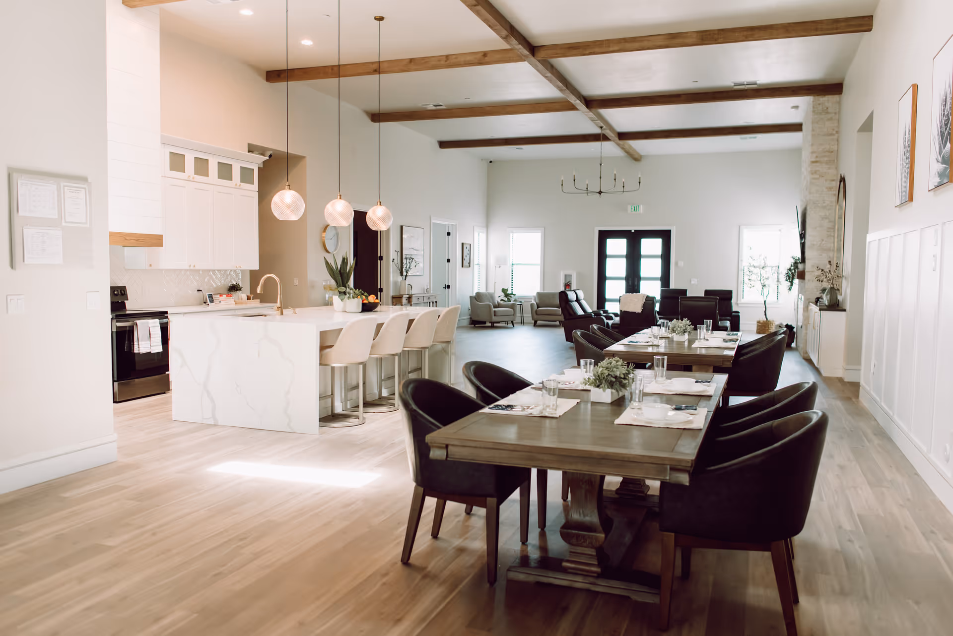 Bright open communal dining and kitchen area with wooden tables, upholstered chairs, a marble island, and exposed ceiling beams.