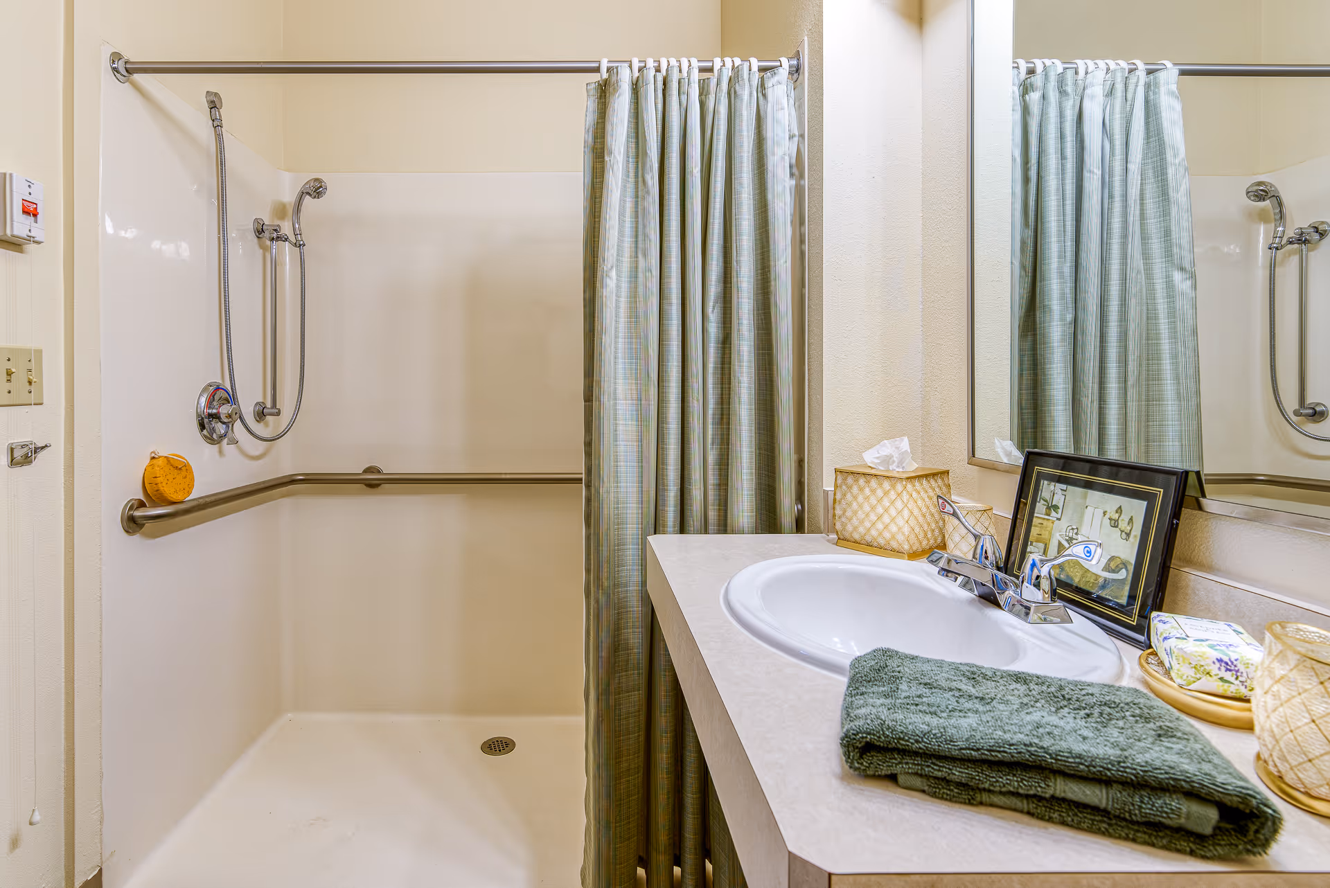 A bathroom with a walk-in shower featuring a handheld showerhead and grab bars. A shower curtain is partially closed. Next to the shower is a countertop with a sink, a folded green towel, a tissue box, a framed picture, and other small items. A large mirror is mounted on the wall above the sink.