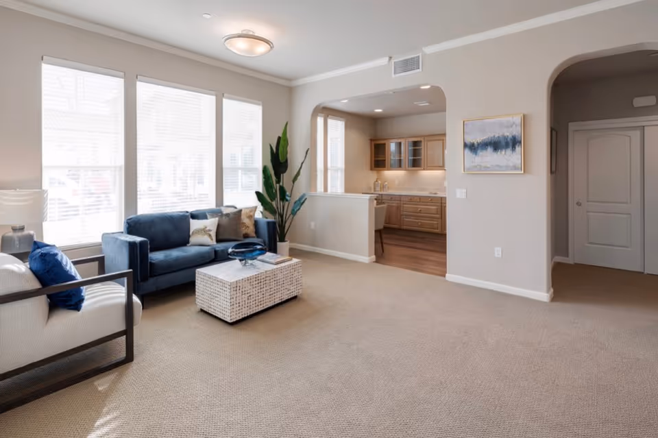 A bright living room with beige carpet, a blue sofa with decorative pillows, a white armchair with a blue pillow, a white woven coffee table, a tall green plant, and large windows with blinds letting in natural light. The room opens into a kitchen area with wooden cabinets and a small dining space. A framed abstract painting hangs on the wall near an arched doorway.