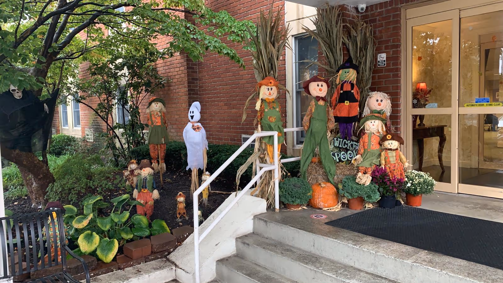 Entrance of a senior living facility decorated with Halloween scarecrows, pumpkins, and a ghost by the front steps.