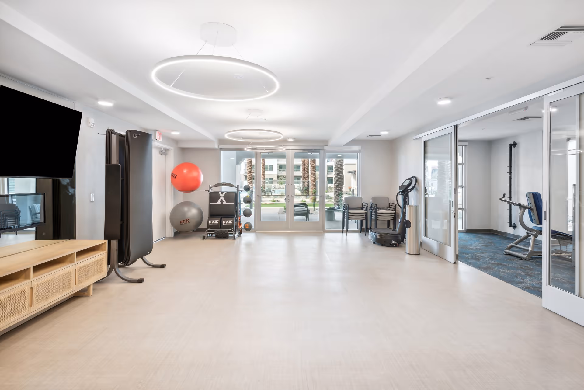 A bright and spacious fitness room with light-colored flooring and modern circular ceiling lights. The room contains exercise equipment including large exercise balls, a rack with smaller balls, stacked chairs, and a vibration plate machine. There is a large flat-screen TV mounted on the wall above a wooden cabinet. Glass doors lead to an adjacent room with additional exercise equipment and windows showing an outdoor area with palm trees and buildings.
