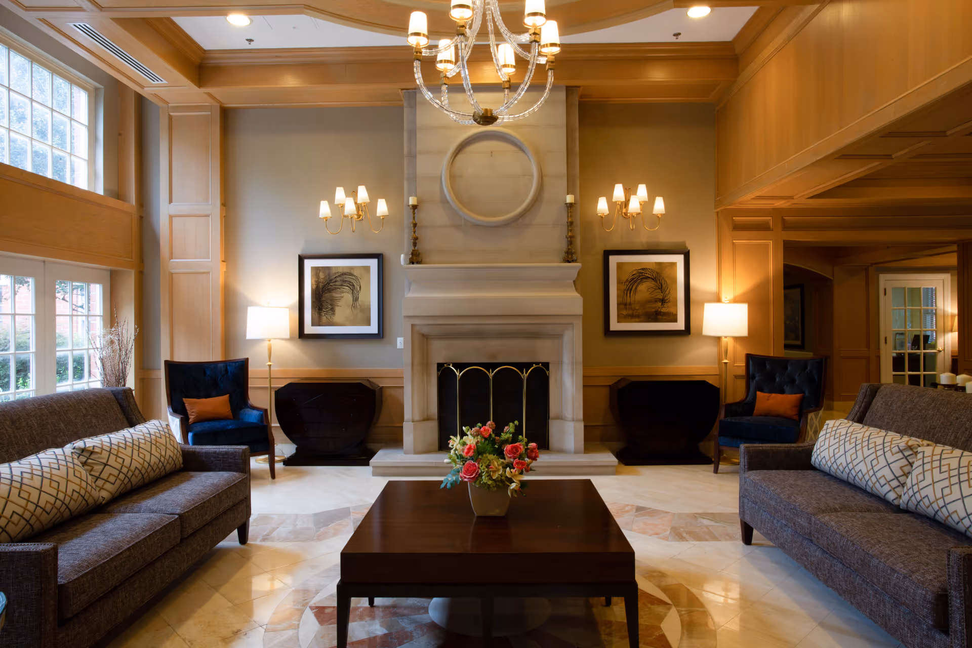 A cozy living room with two gray sofas facing each other, each with patterned cushions. Between them is a dark wooden coffee table with a floral arrangement. The room features a large stone fireplace with a circular decorative element above it, flanked by two framed artworks and wall sconces. Two blue armchairs with orange cushions sit near the fireplace, and large windows on the left allow natural light to fill the space. The ceiling has wooden beams and a chandelier hanging in the center.