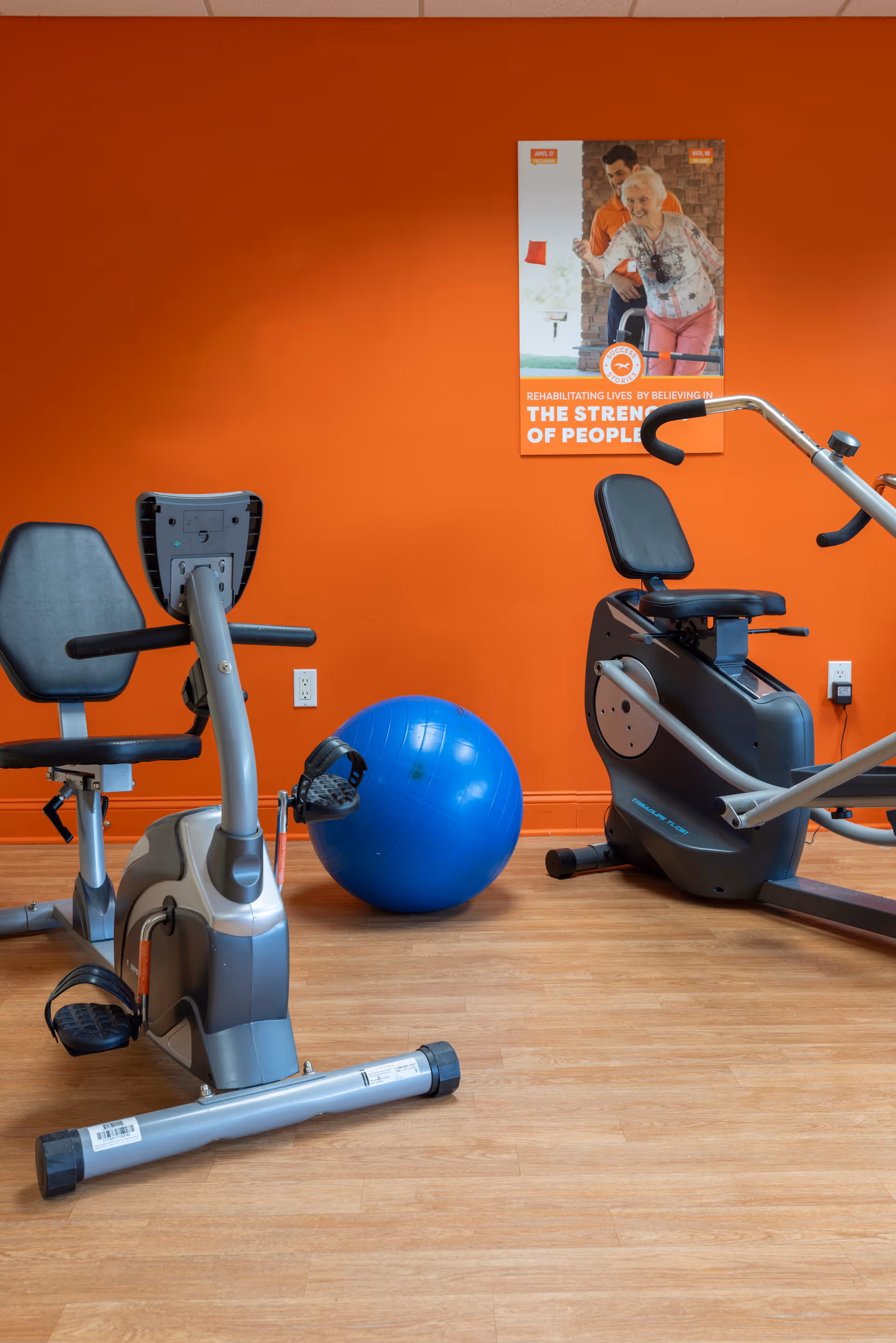 A small exercise room with two stationary recumbent bikes and a blue exercise ball on a wooden floor. The wall behind is painted orange and has a poster showing an elderly woman and a man with text about rehabilitating lives.