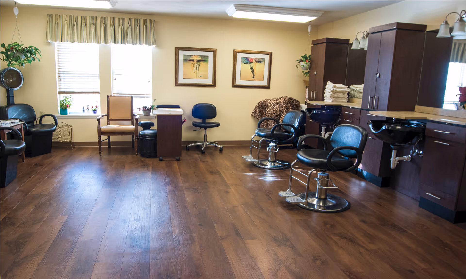 Interior of a salon room with wooden flooring, two black salon chairs in front of black wash basins, a beige armchair, a black rolling stool, a hair dryer chair, plants, and framed artwork on the wall.