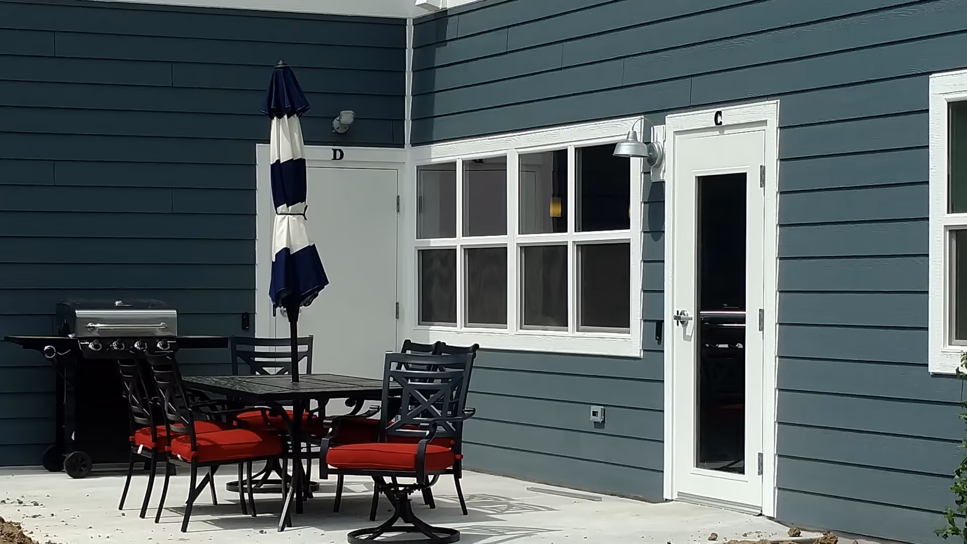 Outdoor patio area with a black metal table and six chairs with red cushions. A closed blue and white striped umbrella is positioned in the center of the table. There is a black grill on the left side near a white door labeled 'D'. Another white door labeled 'C' with a glass panel is on the right side, along with a window with white trim. The building exterior is painted blue with white trim.