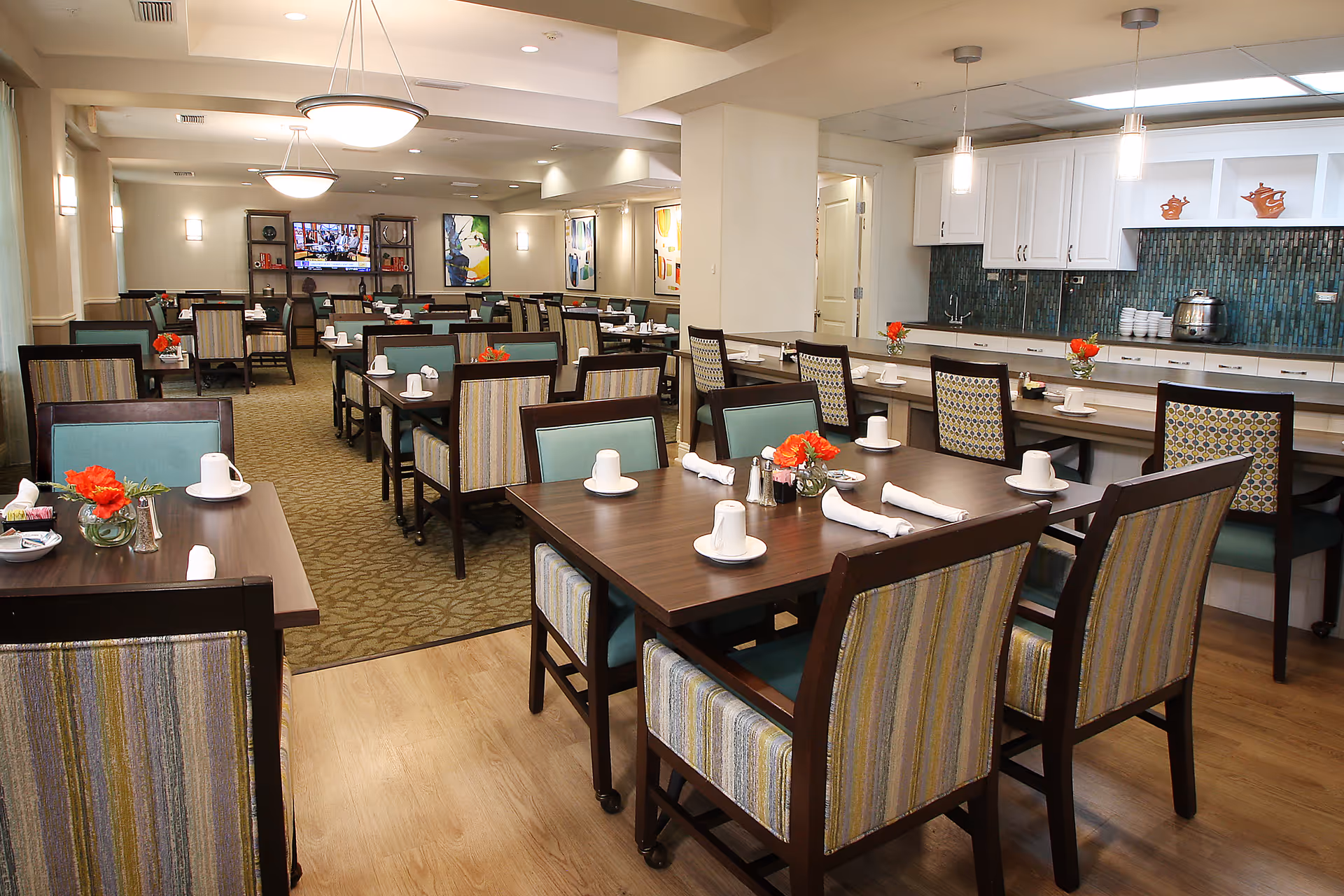 A spacious dining room in Prestonwood Court with multiple tables and chairs arranged neatly. Each table is set with white cups, saucers, napkins, and small flower vases with red flowers. The room features a mix of wood and carpet flooring, modern light fixtures, and a kitchen area with white cabinets and a green tiled backsplash in the background. A television is mounted on the far wall above a shelving unit with decorative items.