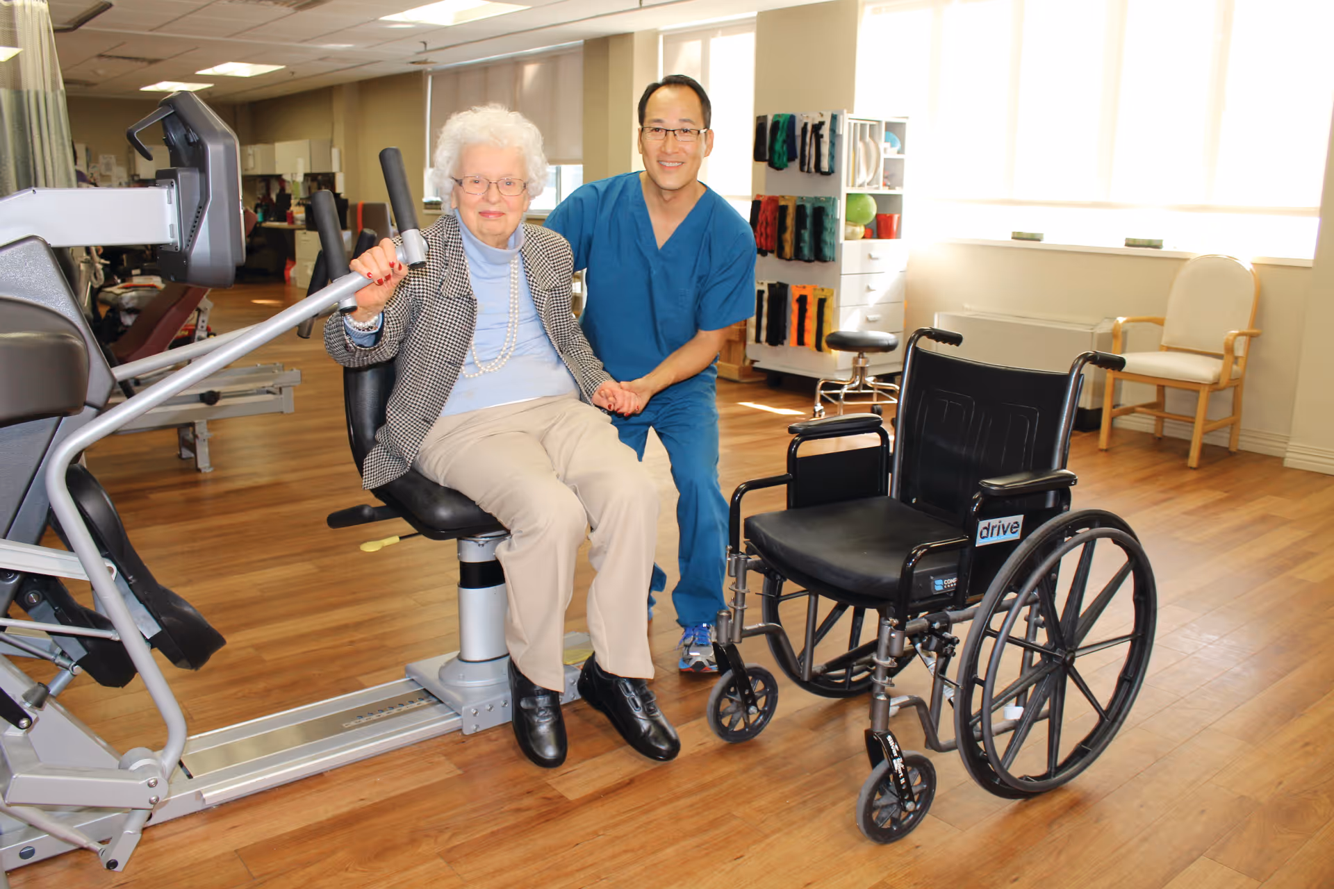 An elderly woman seated on a piece of exercise equipment in a rehabilitation or physical therapy room, holding the hand of a smiling male healthcare professional in blue scrubs kneeling beside her. A wheelchair is positioned nearby, and the room has wooden floors, exercise equipment, and shelves with colorful therapy supplies.
