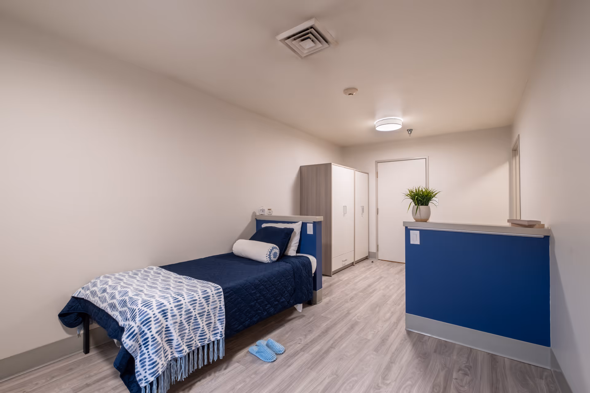 A simple, clean bedroom in a memory care facility featuring a single bed with navy blue bedding and a patterned throw blanket. There is a white and wood wardrobe against the wall, a small blue half-wall with a potted plant on top, and light wood flooring. The room has plain white walls and a ceiling light.