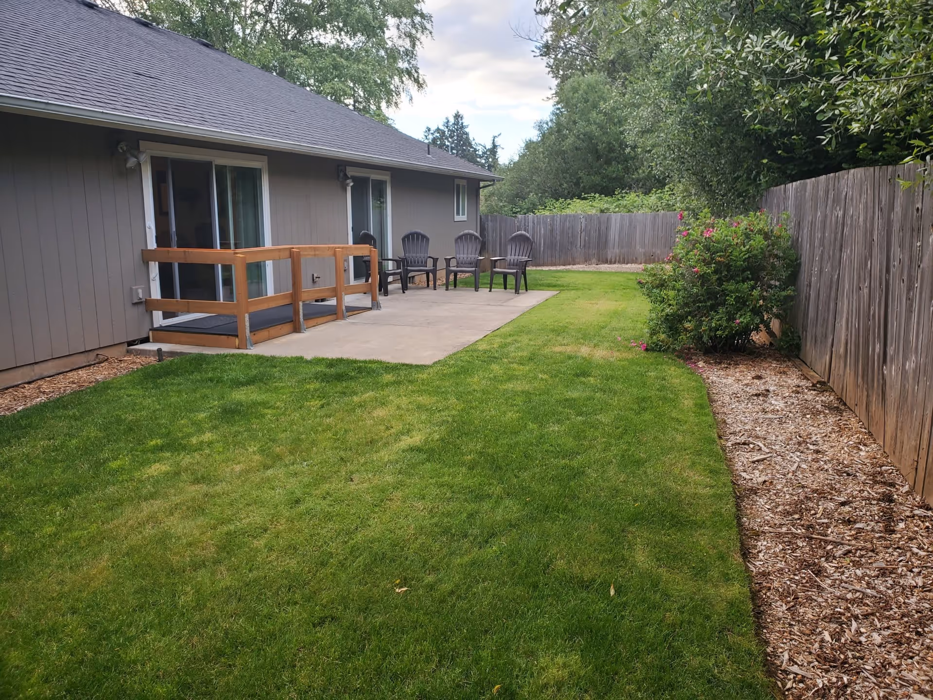 A backyard area of a facility with a green lawn, a concrete patio with four black chairs, a wooden railing near sliding glass doors, a wooden fence enclosing the yard, and some bushes along the fence.