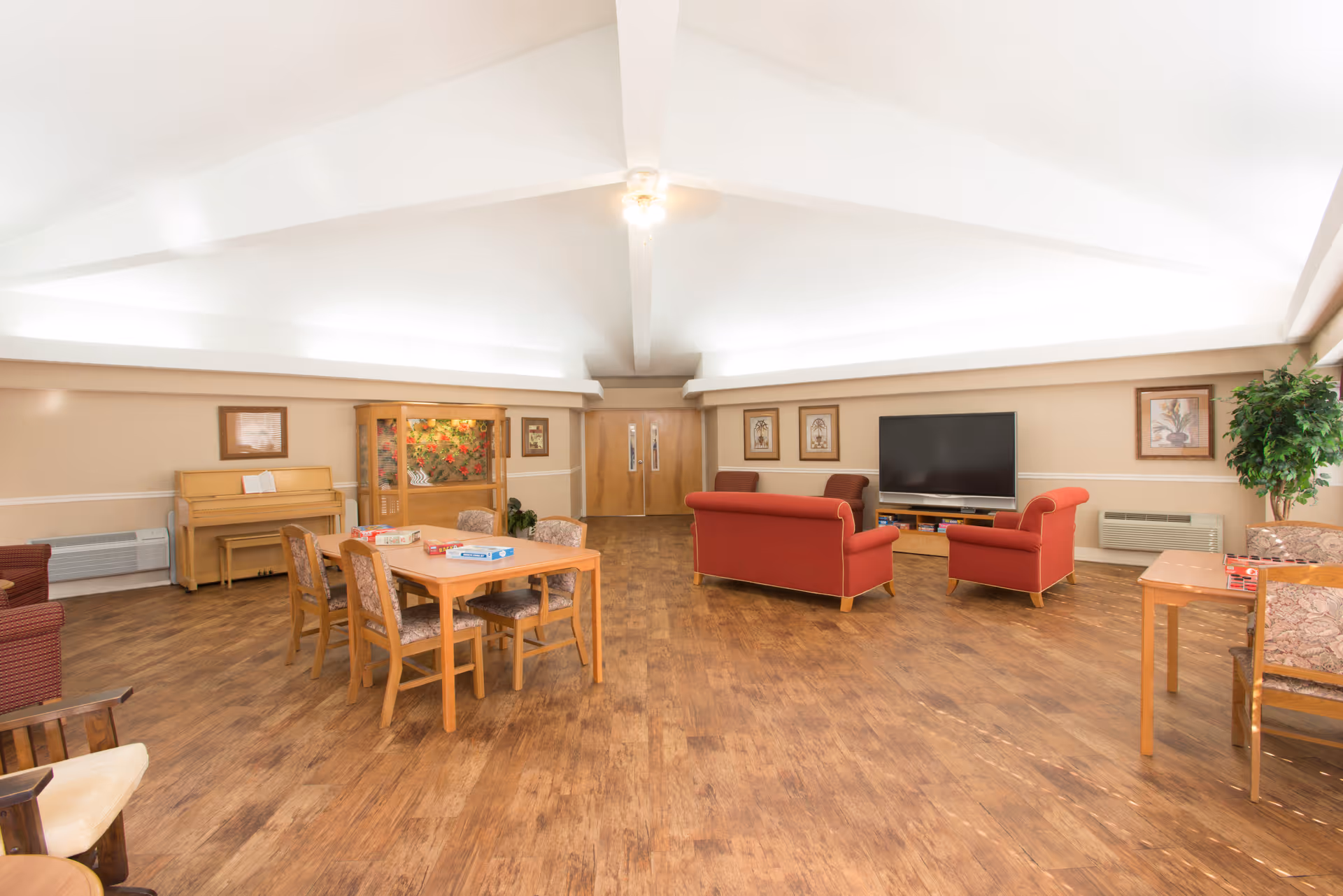 A spacious senior living common area with wooden flooring, a table with four chairs, a red sofa set facing a large flat-screen TV, a piano against the wall, framed artwork, and a potted plant in the corner.