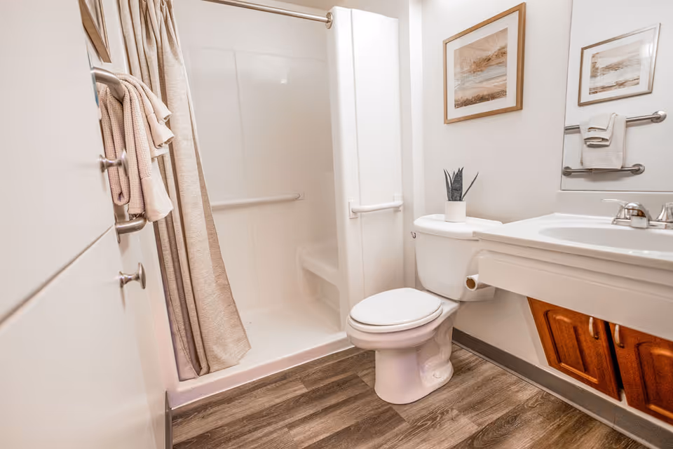 A clean and modern bathroom featuring a white toilet, a white sink with wooden cabinet doors underneath, a walk-in shower with a beige curtain, and a small potted plant on the toilet tank. The walls are white with two framed pictures above the toilet, and there are towels hanging on a bar next to the sink and on a hook near the shower.