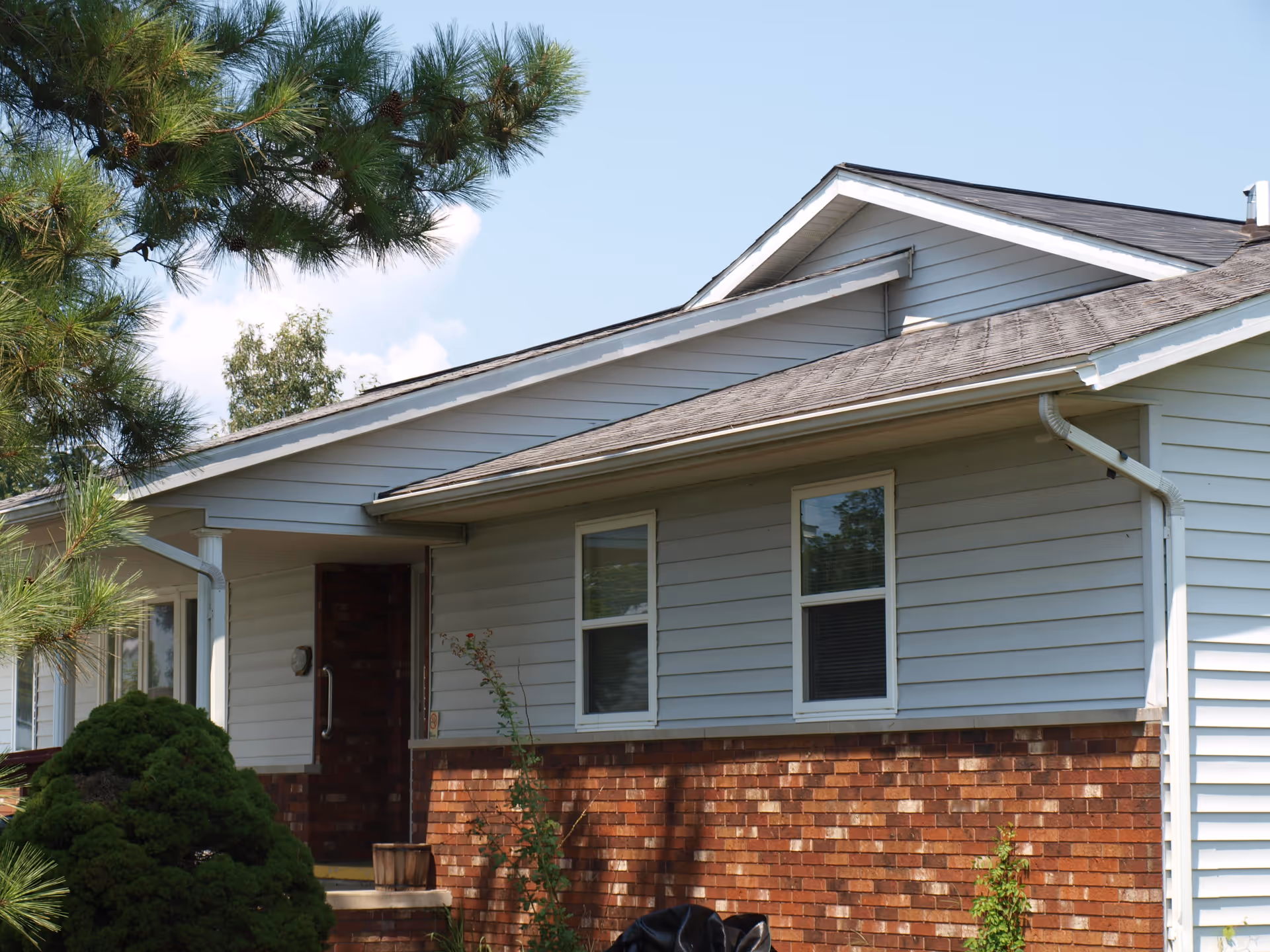 Single-story house front with a brick lower facade, white siding, two windows, a small covered porch, and surrounding shrubs and trees.