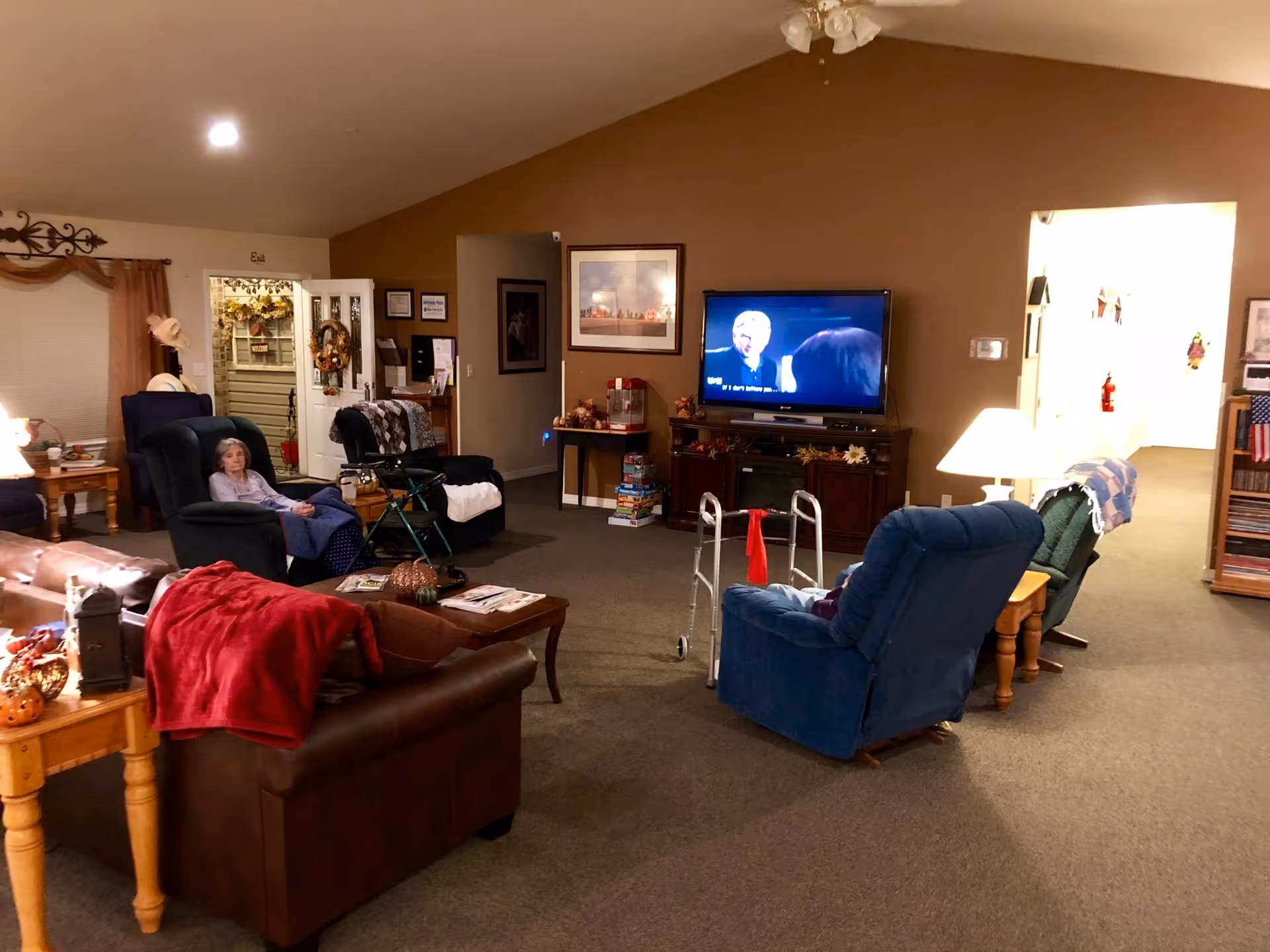 A cozy assisted living facility common room with several armchairs and a sofa arranged around a TV mounted on a wooden cabinet. An elderly woman is sitting in one of the armchairs. There is a walker near another chair, a side table with a lamp, and various decorations including a wreath on the door and framed pictures on the walls. The room has warm lighting and carpeted floors.