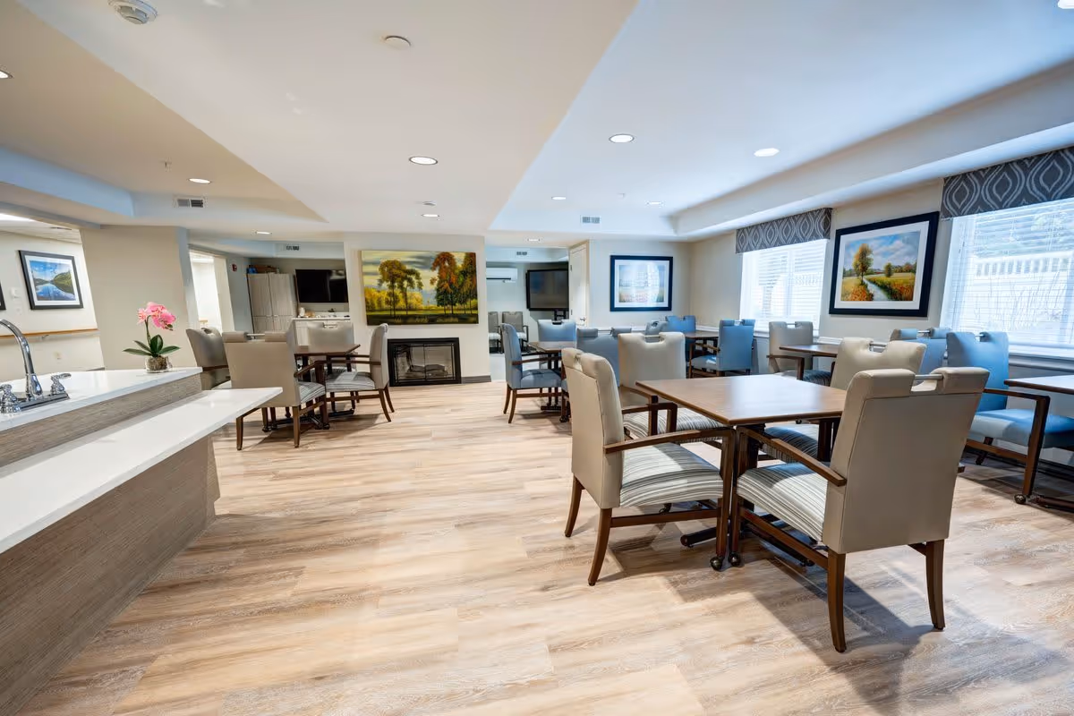 A bright and spacious dining room in a senior living facility with multiple tables and cushioned chairs arranged neatly. The room features large windows with patterned valances, framed landscape paintings on the walls, a fireplace, and a countertop with a sink and a pink orchid plant. The flooring is light wood, and the ceiling has recessed lighting.