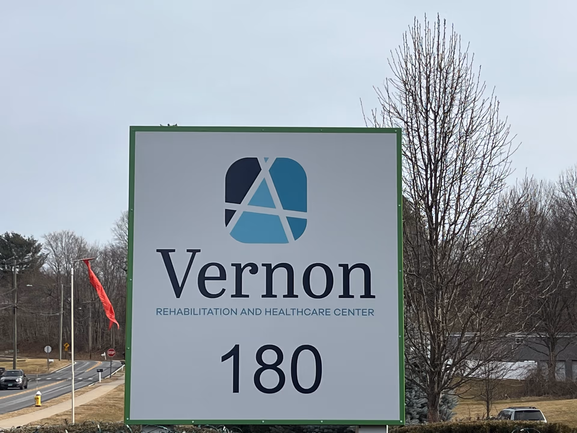 Outdoor sign for Vernon Rehabilitation and Healthcare Center showing the facility logo and the number 180 with trees and a road in the background.