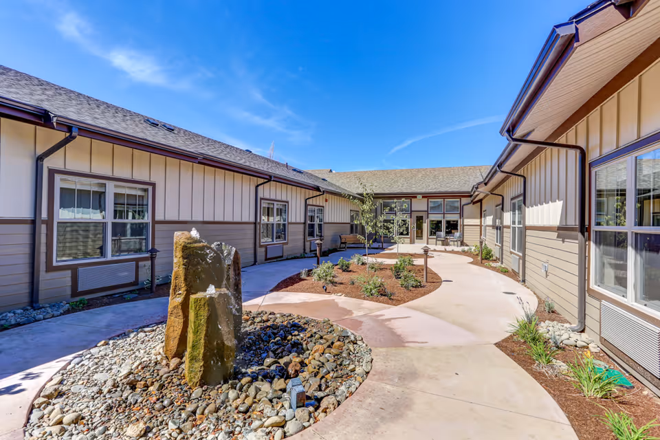 Sunny landscaped central courtyard with a stone water feature, pathways, and surrounding single-story memory care building.