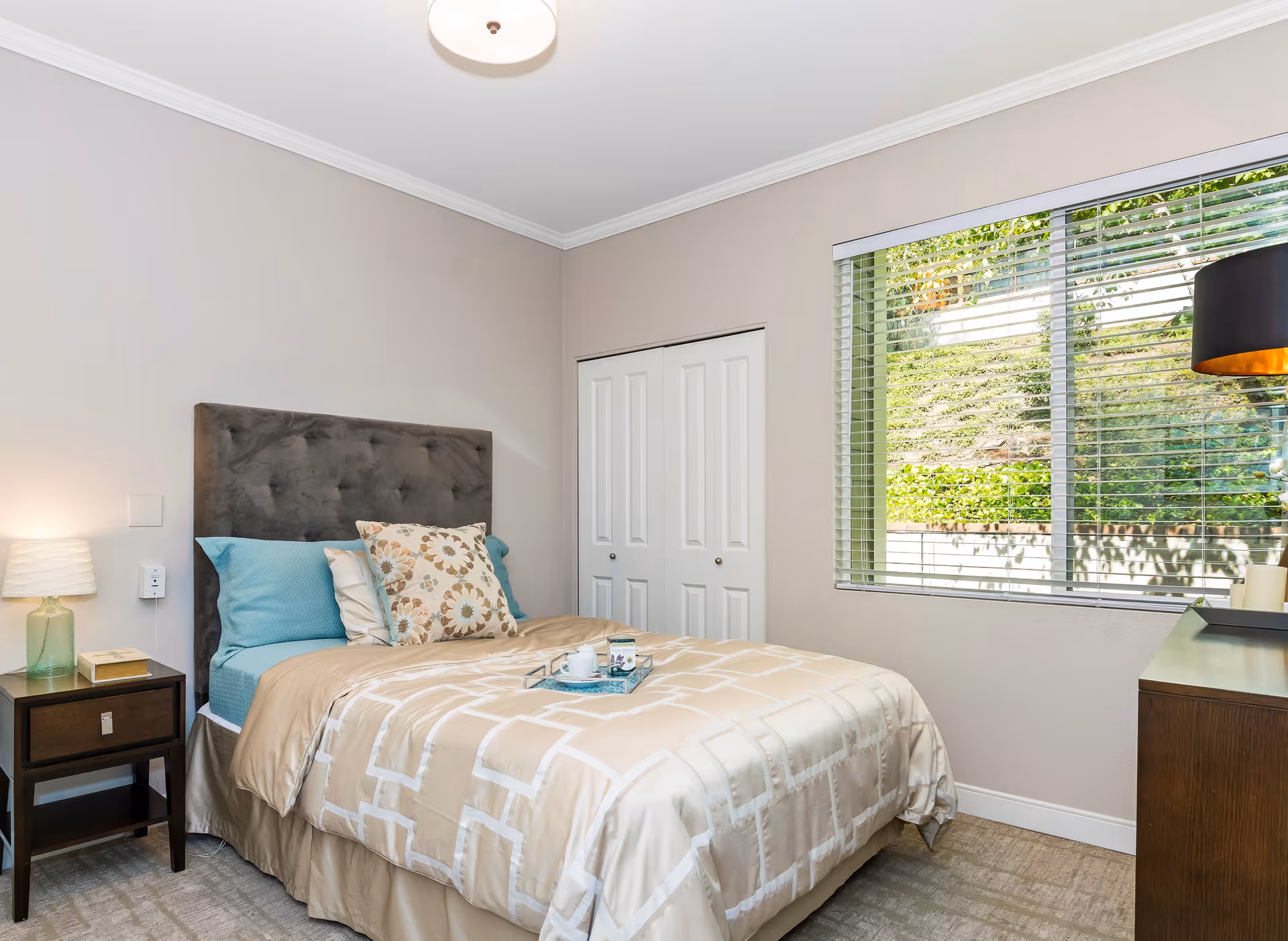 A cozy bedroom with a neatly made bed featuring beige and blue bedding, a dark upholstered headboard, and decorative pillows. There is a wooden nightstand with a lamp and a book on the left side of the bed. A window with blinds shows greenery outside, and a wooden dresser with a black lamp is on the right side of the room. The walls are painted light beige and the floor is carpeted.