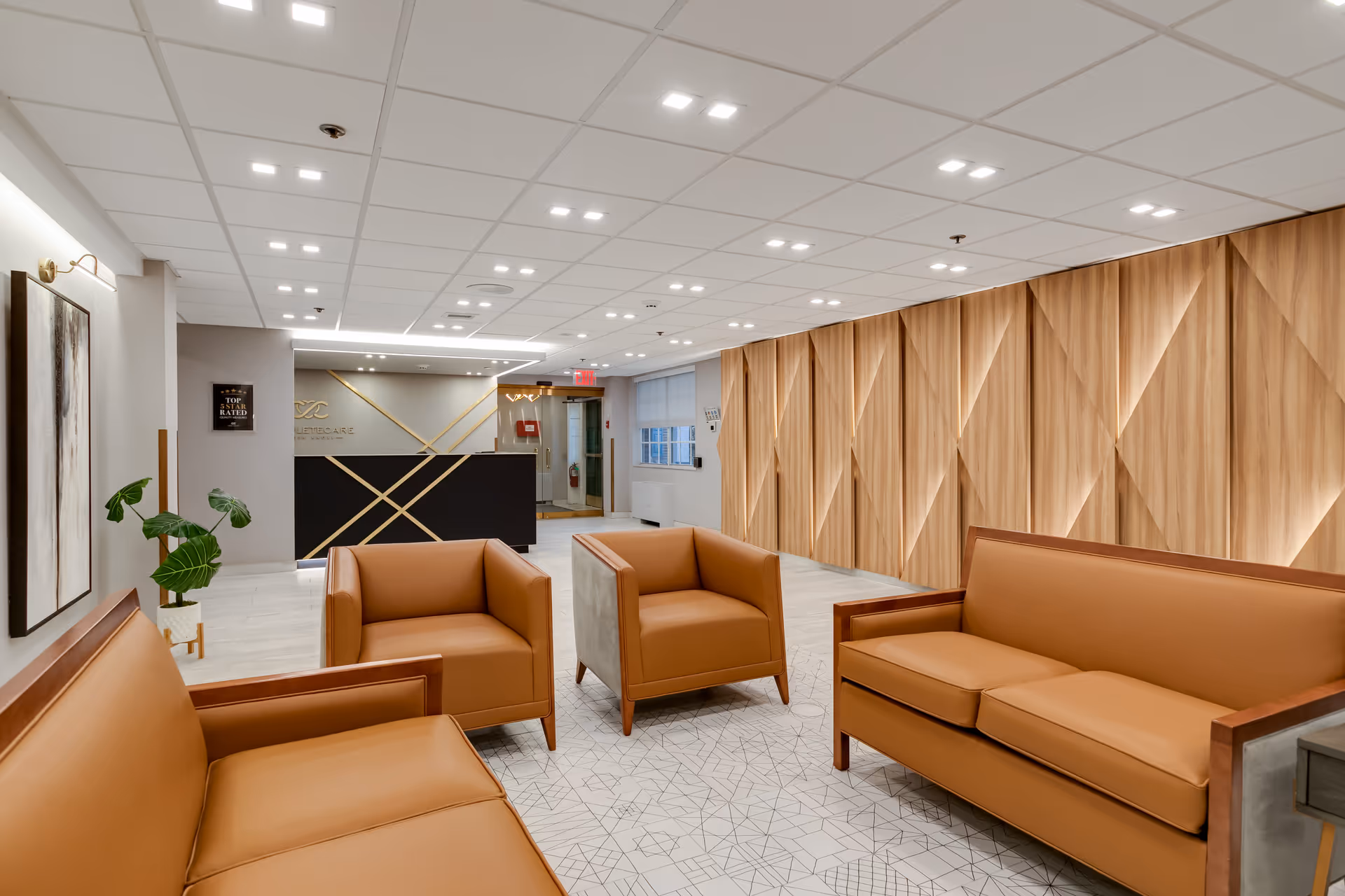 A modern and stylish waiting area with tan leather sofas and armchairs arranged on a patterned tile floor. The back wall features a geometric wooden panel design, and there is a reception desk with gold accents in the background. The ceiling has recessed lighting, and a potted plant and framed artwork decorate the space.