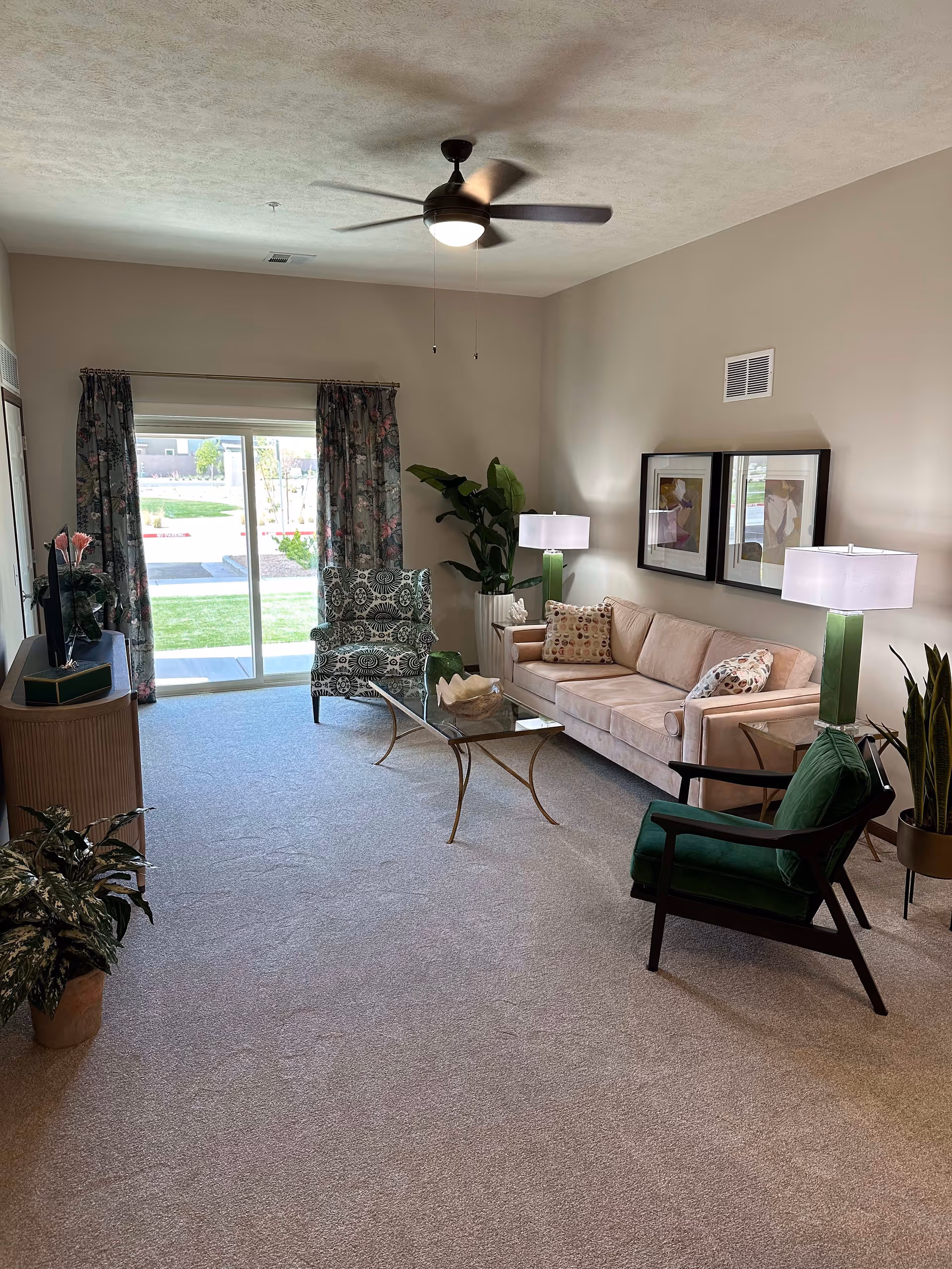 A bright living room with beige walls and carpet. The room features a beige sofa with patterned pillows, a green armchair, and a patterned armchair. There is a glass coffee table with a decorative bowl in the center. Two framed artworks hang on the wall above the sofa. Two table lamps with green bases and white shades are placed on either side of the sofa. A ceiling fan with a light is mounted on the ceiling. Sliding glass doors with floral curtains lead to an outdoor area. Several potted plants are placed around the room.