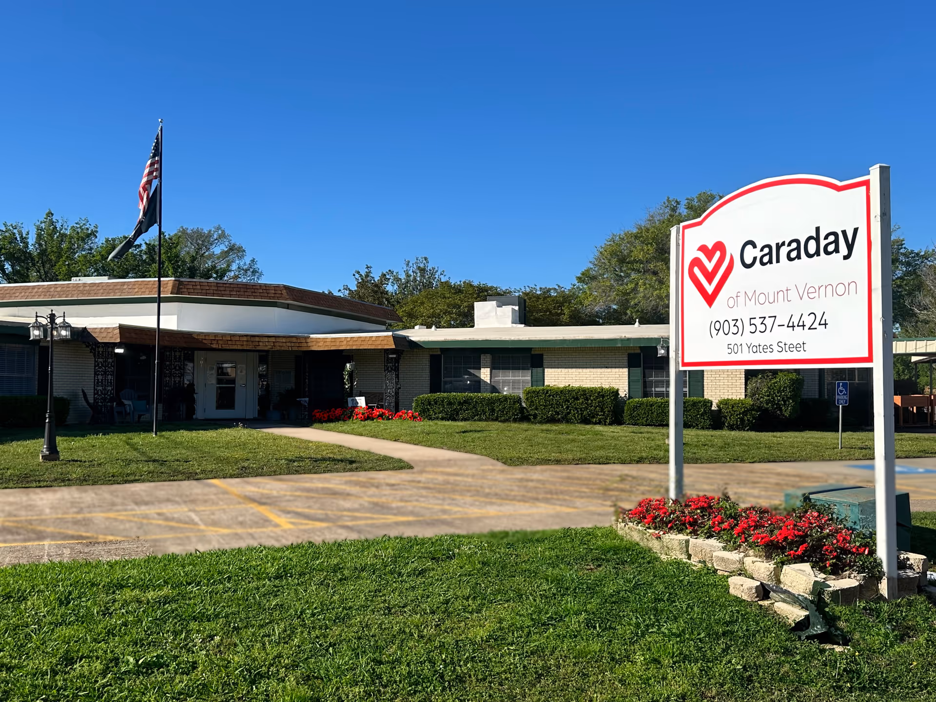 Front exterior of Caraday of Mount Vernon with a lawn, flagpole, flowerbeds, and a large sign showing the facility name and phone number.