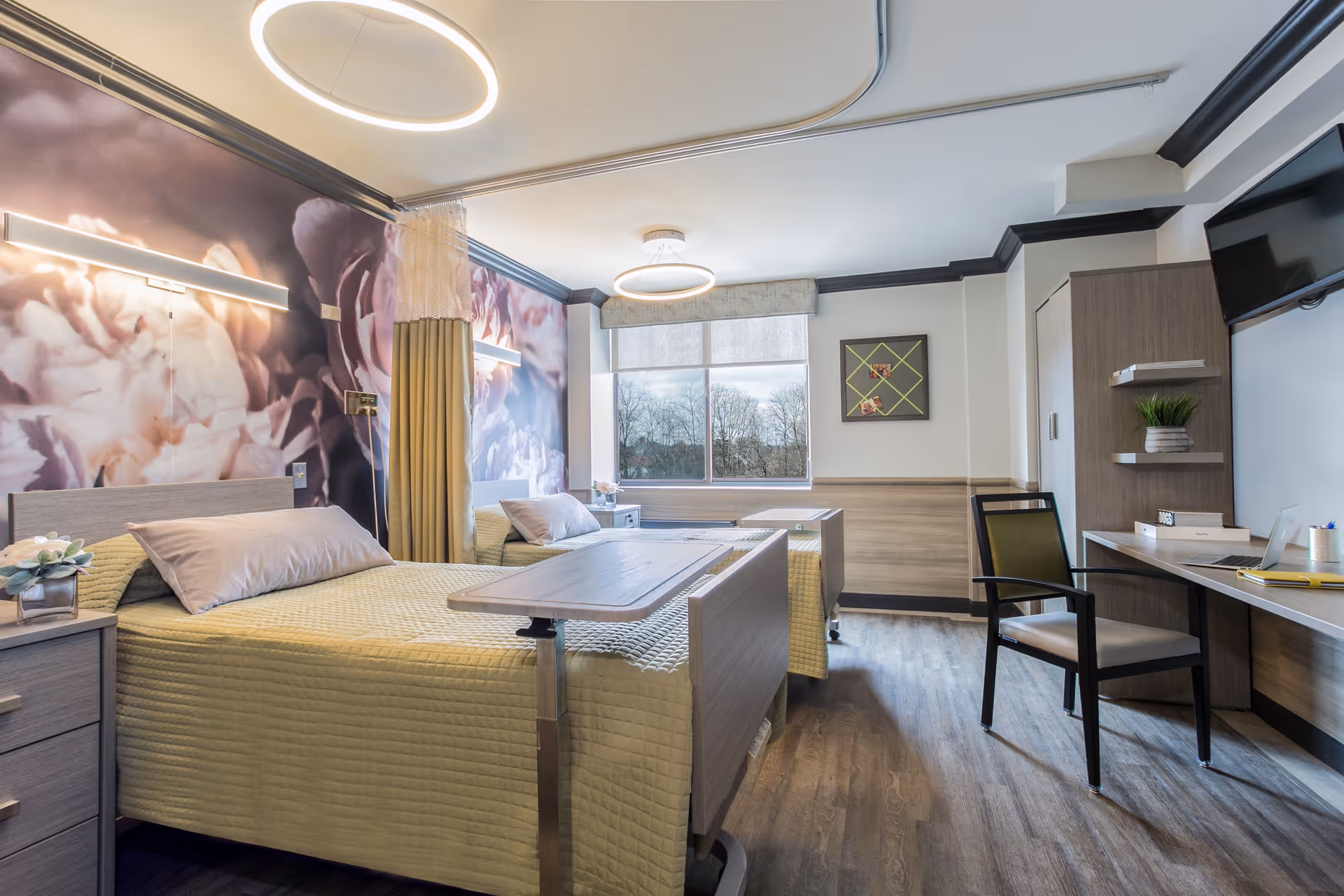 A bright and modern rehabilitation and nursing room with two hospital-style beds, each with beige bedding and pillows. The room features a large window with a view of trees outside, a floral mural on one wall, a curtain divider between the beds, a desk with a chair, a mounted TV, and wooden flooring.