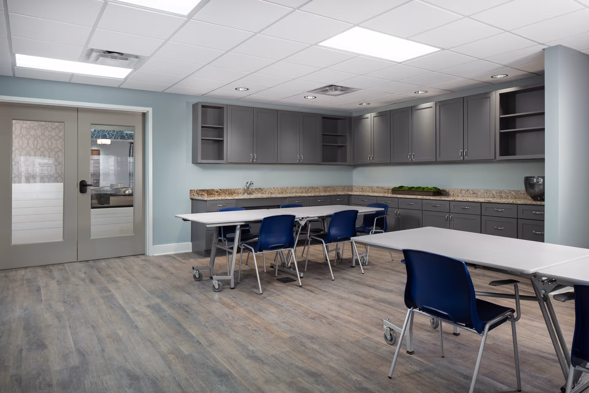 A spacious room with light blue walls and wood-look flooring featuring two white tables with blue chairs on wheels. The back wall has gray cabinets with granite countertops and a sink. Double glass doors are visible on the left side of the room.