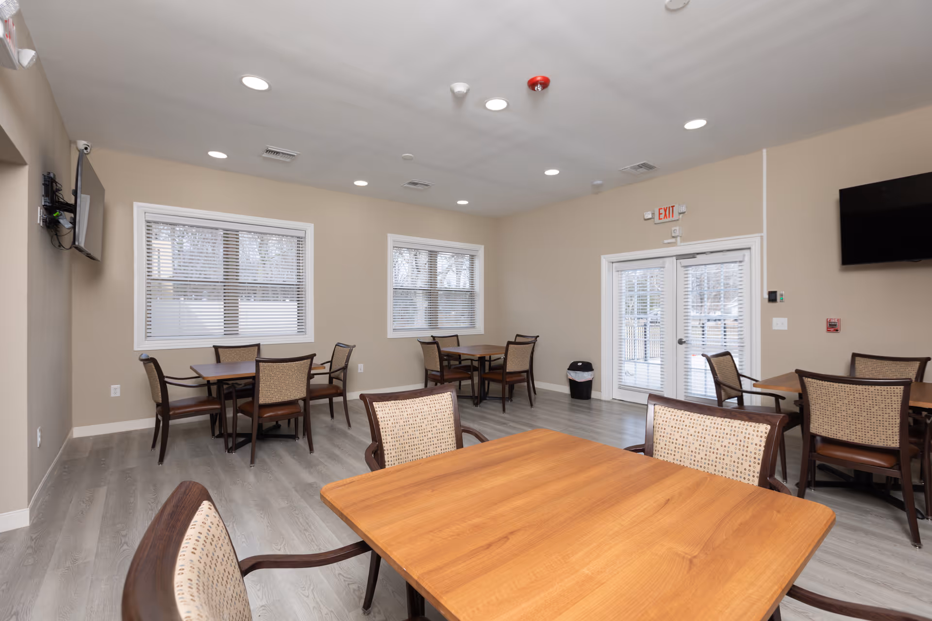 A clean and well-lit dining room with several wooden tables and cushioned chairs arranged neatly. The room has light-colored walls, large windows with blinds, a wall-mounted TV, and double glass doors leading outside. The floor is covered with light gray wood-like flooring.