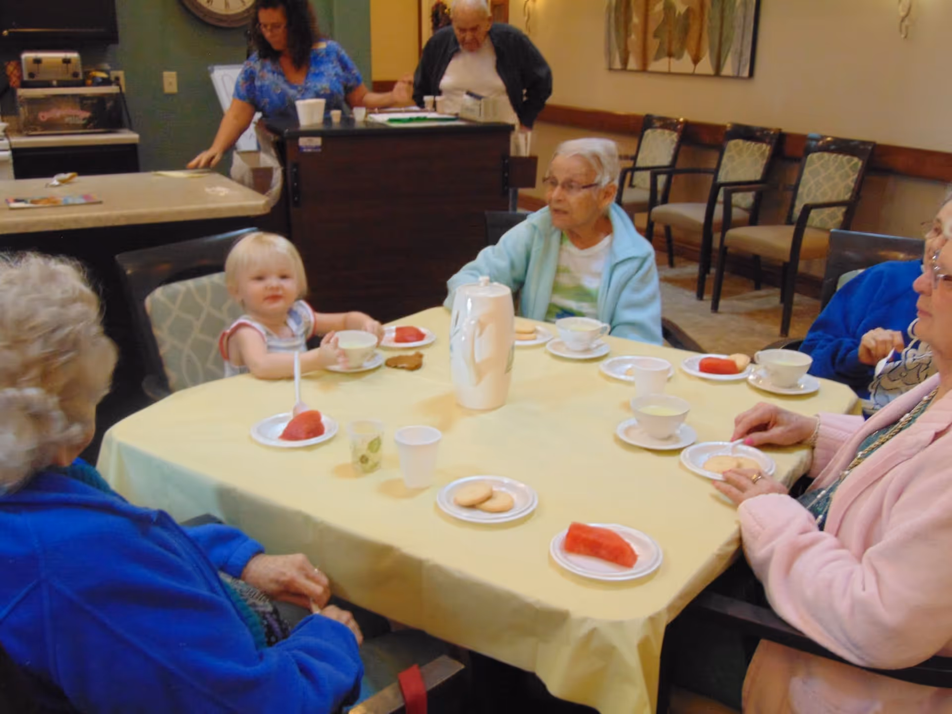 A group of residents and a toddler sit around a table in a communal dining room with teacups, cookies, and slices of watermelon.