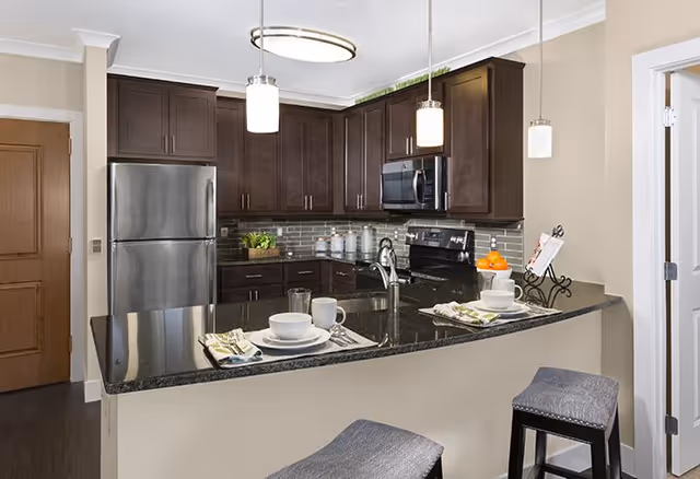 Modern kitchen with dark wood cabinets, stainless steel refrigerator and microwave, black granite countertop with two place settings, two gray cushioned bar stools, and pendant lights hanging above the counter.