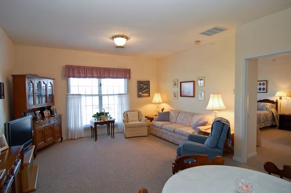 Bright living room with a sofa, armchairs, wooden cabinets, TV and a large window opening to a bedroom.